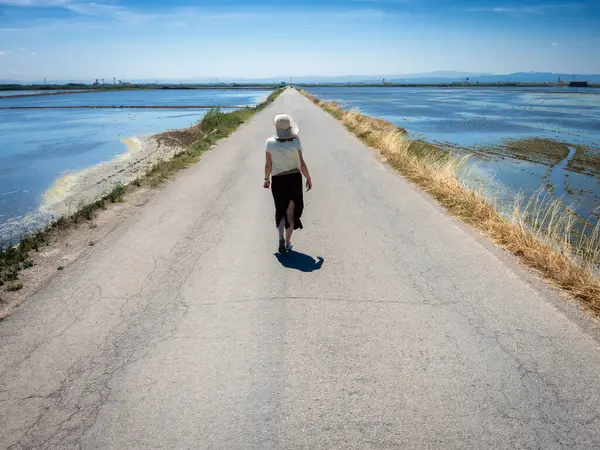 Şapkalı bir kadın ıssız bir yolda yürüyor, kenarlarında su var.