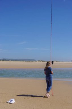 Deniz kıyısında çıplak ayaklı bir kadın, elinde bir olta kamışı, bulutsuz mavi bir gökyüzünün altında