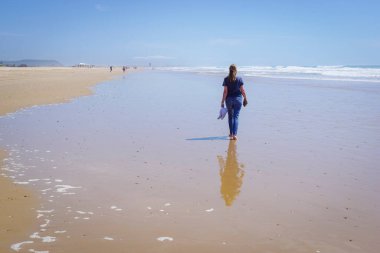 Yalınayak bir kadın, arka planda mavi deniz olan uzun, altın bir kumsal kıyısında yürüyor.