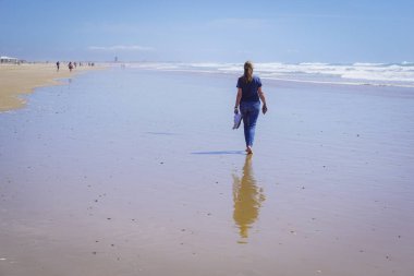 Yalınayak bir kadın, arka planda mavi deniz olan uzun, altın bir kumsal kıyısında yürüyor.