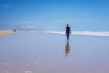 Yalınayak bir kadın, arka planda mavi deniz olan uzun, altın bir kumsal kıyısında yürüyor.