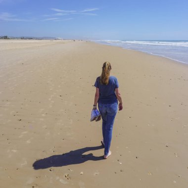 Yalınayak bir kadın, arka planda mavi deniz olan uzun, altın bir kumsal kıyısında yürüyor.