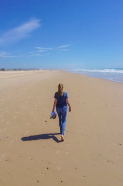 Yalınayak bir kadın, arka planda mavi deniz olan uzun, altın bir kumsal kıyısında yürüyor.