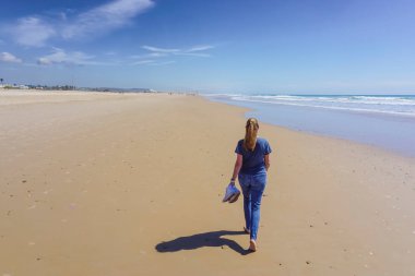 Yalınayak bir kadın, arka planda mavi deniz olan uzun, altın bir kumsal kıyısında yürüyor.
