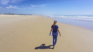 Yalınayak bir kadın, arka planda mavi deniz olan uzun, altın bir kumsal kıyısında yürüyor.