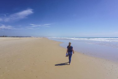 Yalınayak bir kadın, arka planda mavi deniz olan uzun, altın bir kumsal kıyısında yürüyor.