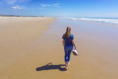 Yalınayak bir kadın, arka planda mavi deniz olan uzun, altın bir kumsal kıyısında yürüyor.