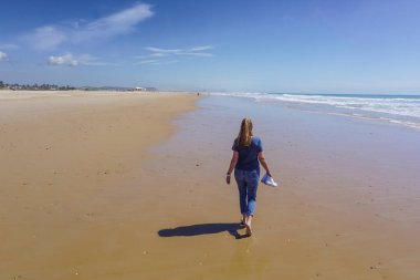 Yalınayak bir kadın, arka planda mavi deniz olan uzun, altın bir kumsal kıyısında yürüyor.