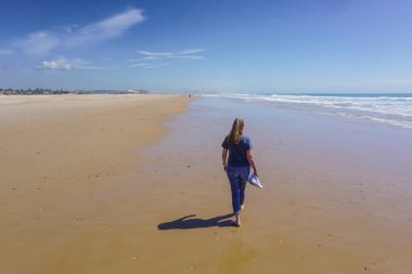 Yalınayak bir kadın, arka planda mavi deniz olan uzun, altın bir kumsal kıyısında yürüyor.