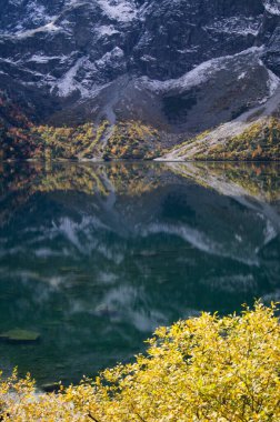 Büyüleyici Morskie Oko Gölü karla kaplı Tatra Dağları 'na karşı sonbahar paletini yansıtıyor. Seyahat, mevsimsel değişim ve Doğu Avrupa manzaralarının güzelliği üzerine makaleler için mükemmel..