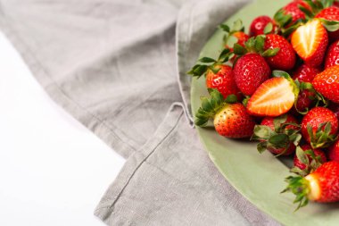 Açık yeşil tabakta kırmızı çilekler ve beyaz arka planda gri keten peçeteler..