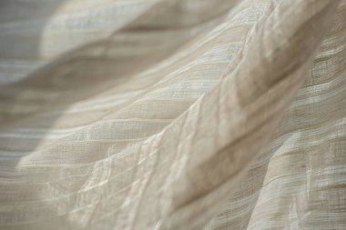 Işık ışınlarının arka planına karşı bej bej renginde şeffaf kumaş. Pencerenin arka planında bir şeritte ışık kumaşı katları.