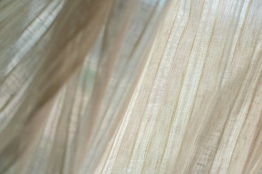 Işık ışınlarının arka planına karşı bej bej renginde şeffaf kumaş. Pencerenin arka planında bir şeritte ışık kumaşı katları.