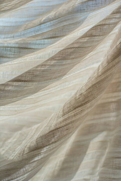 Light linen transparent fabric in beige against the background of light rays. Folds of light fabric in a strip on the background of the window.