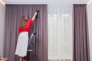 Koyu bej perdeleri el yapımı buharlı gemiyle ütüleyen kadın..