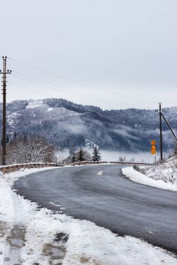 Sis ve mavi gökyüzüyle dağlara giden yol. Avrupa 'da kış. Ağaçlar ve yol boyunca uzanan karlar ormana düştü. Yol yılanlarla dolu. Doğanın güzel bir manzarası. Otobanda yol işaretleri var. Çelik yol çitleri. İletişim kabloları.