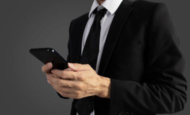 Siyah takım elbiseli bir işadamı iş verilerini görmek ve işi kontrol etmek için elinde cep telefonu tutuyor.