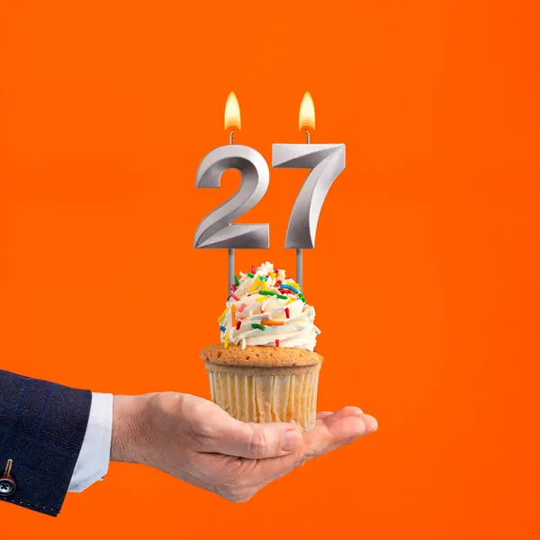 The hand that delivers cupcake with the number 27 candle - Birthday on orange background