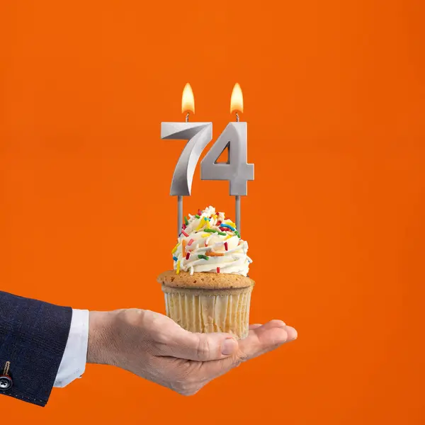 The hand that delivers cupcake with the number 74 candle - Birthday on orange background
