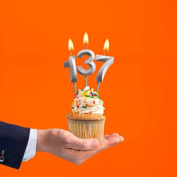 stock image Hand holding birthday cupcake with number 137 candle - background orange