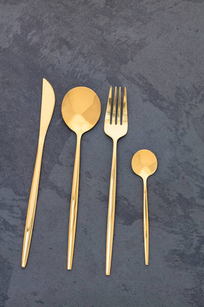 Элегантные золотые столовые приборы. Вилка, нож и ложки на темном каменном фоне.