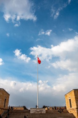 Üzerinde Türk bayrağı olan uzun bir bayrak direği, kısmen bulutlu, canlı mavi bir gökyüzüne gururla el sallıyor. Bayrak büyük bir taş heykelin önünde dikiliyor ve insanlar merdiven çıkıyor..