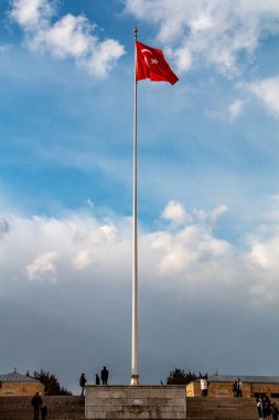 Uzun bir bayrak direği gururla duruyor ve bulutlu bir gökyüzünün zemininde Türk bayrağını sergiliyor. Uzakta, bayrak direğinin tabanında birkaç birey görülüyor ve olay yerine bir ölçek hissi ekleniyor. Genel kompozisyonu bir...