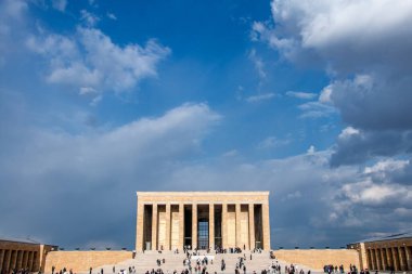 Görüntüde Ankara, Türkiye 'deki Antkabir, Atatrk' ın Mozolesi yer alıyor. Görkemli bej renkli taş cephesi, detaylı korniş ve tek, büyük, yansıtıcı merkezi bir kapı, parlak mavi bir gökyüzüne karşı belirgin bir şekilde sergilenmektedir. Bir güvenlik. 