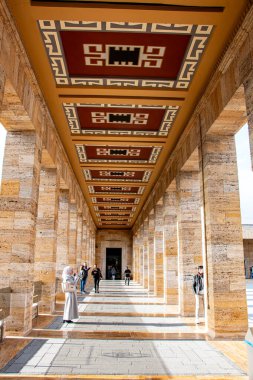 Açık bej renkli taş sütunlardan oluşan uzun bir koridora, turuncu, kırmızı ve gri tonlarında tekrarlanan geometrik tasarımlara sahip desenli bir tavana ve uzayda yürüyen birkaç kişiye bakış açısı.