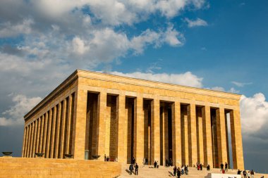 Görüntüde Ankara, Türkiye 'deki Antkabir, Atatrk' ın Mozolesi yer alıyor. Görkemli bej renkli taş cephesi, detaylı korniş ve tek, büyük, yansıtıcı merkezi bir kapı, parlak mavi bir gökyüzüne karşı belirgin bir şekilde sergilenmektedir. Bir güvenlik. 