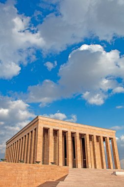 Görüntüde Ankara, Türkiye 'deki Antkabir, Atatrk' ın Mozolesi yer alıyor. Görkemli bej renkli taş cephesi, detaylı korniş ve tek, büyük, yansıtıcı merkezi bir kapı, parlak mavi bir gökyüzüne karşı belirgin bir şekilde sergilenmektedir. Bir güvenlik. 