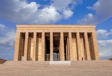 Görüntüde Ankara, Türkiye 'deki Antkabir, Atatrk' ın Mozolesi yer alıyor. Görkemli bej renkli taş cephesi, detaylı korniş ve tek, büyük, yansıtıcı merkezi bir kapı, parlak mavi bir gökyüzüne karşı belirgin bir şekilde sergilenmektedir. Bir güvenlik. 