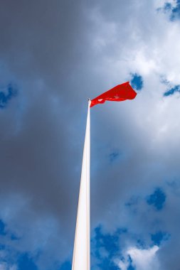 Düşük açılı tek bir kırmızı bayrak uzun, ince beyaz bir direkte parçalı bulutlu mavi bir gökyüzünün zemininde dalgalanıyor. Görüntü dikey odaklıdır, kutbun yüksekliğini ve gökyüzünün genişliğini vurgular..