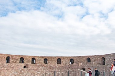 Parçalı bulutlu bir gökyüzünün altındaki tarihi taş kale duvarının panoramik görüntüsü. Duvarın çevresinde birkaç kişinin yürüdüğü görülüyor..