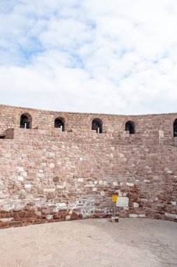 Görüntü, parçalı bulutlu bir gökyüzünün altında kadim bir taş duvarın kıvrımlarını gösteriyor. Duvar çeşitli boyutlarda kırmızımsı-kahverengi taşlardan yapılmıştır ve çeşitli kemerli açıklıklara sahiptir. W 'nin tabanındaki asfalt alanda küçük, sarı bir tabela duruyor.