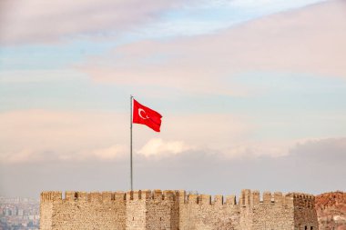 Canlı kırmızı bir Türk bayrağı yumuşak, pastel bir gökyüzüne hafifçe dalgalanır, gururla yıpranmış antik taş kale duvarlarının üzerinde durur. Sahne, tarih ve ulusal gurur duygusunu çağrıştırıyor..