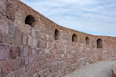 Eski taş bir duvarın kıvrımlı bir bölümü yıpranmış dokusunu ve sağlam yapısını sergiliyor. Ruh hali, tarihsel öneme ve sessiz kuvvete sahiptir. Dekor açık havada, parçalı bulutlu bir gökyüzünün altında..