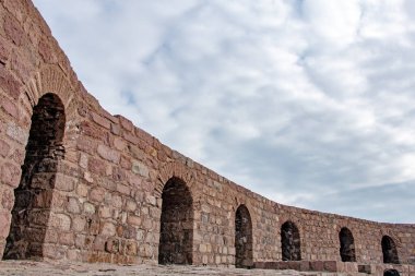 Görüntü, parçalı bulutlu bir gökyüzü altında yavaşça kıvrılan antik bir taş su kemerinin bir bölümünü gösteriyor. Yapı, titizlikle düzenlenmiş kırmızımsı kahverengi taşlardan yapılmıştır. Su kemerinin uzunluğu boyunca sayısız kemerli açıklıklar görülebilir. 