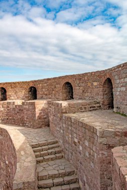 Bir fotoğraf, kadim taş duvarların kemerli delikli bir bölümünü gösteriyor. Yapının içinden geçen taş bir patika ve merdiven kalenin içine bir göz atmamızı sağlıyor. Gökyüzü kısmen görülebilir, bulutlar ve mavi karışımı ile.