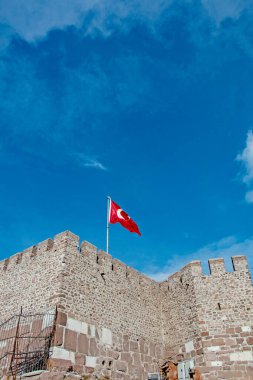 Antik bir taş kale duvarının tepesinde canlı mavi bir gökyüzüne karşı kırmızı bir Türk bayrağı dalgalanır..
