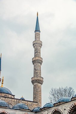 İstanbul 'daki bir caminin uzun ve ince minaresinin yakın plan görüntüsü, karmaşık taş işçiliği ve sessiz gökyüzüne karşı sivri bir kule. Resmin alt kısmında cami çatılarının çeşitli kubbeleri olan kısmı görülebiliyor..