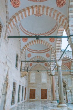 Bir caminin içinin nefes kesici bir perspektifi. Çizgili kemerler ve karmaşık tavan detayları büyüleyici bir ışık ve gölge deseni yaratır. Soluk taş duvarlar ve mermer zemin süslemeli ahşap kapılarla noktalanmış huzurlu atmosferi güçlendirir. 