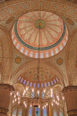 Sultan Ahmed Camii 'nin içinin nefes kesici manzarası, kaligrafi ve canlı renklerle süslenmiş karmaşık kubbesini gözler önüne seriyor. Aşağıda görkemli bir avize asılı, mimarinin ihtişamını artırıyor. Görüntü güzelliği yakalar.