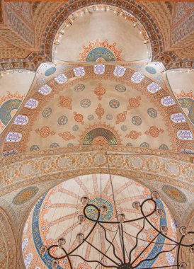 Yukarı doğru nefes kesici bir görüntü Sultan Ahmed Camii 'nin kubbesinin süslü detaylarını yakalıyor. Karmaşık kaligrafi ve renkli desenler aşağıda asılı zarif bir avizeyle tamamlanmış geniş tavanı süsler. Görüntü caminin resmini gösteriyor. 