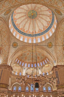 Sultan Ahmed Camii 'nin iç mekanının çarpıcı bir perspektifi. Kubbenin, kemerlerin, el yazısının ve vitray pencerelerinin karmaşık ayrıntıları açıkça görülüyor. Mimari mucizenin merkezinde büyük bir avize asılı..