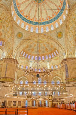 Sultan Ahmed Camii 'nin İstanbul, Türkiye' deki iç manzarası nefes kesici. Görüntü, karmaşık İslam kaligrafisi ve canlı renklerle süslenmiş görkemli kubbeyi sergiliyor. Merkezden sarkan büyük, süslü bir avize onu aydınlatıyor.