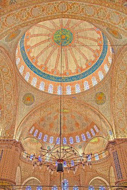 Sultan Ahmed Camii 'nin iç mekanına nefes kesici bir manzara. Görüntü, altta asılı büyük bir avizeyle tamamlanmış, karmaşık kaligrafi ve canlı renklerle süslenmiş çarpıcı kubbeyi sergiliyor. Süslü detaylar ve kemerli yapılar oluştur