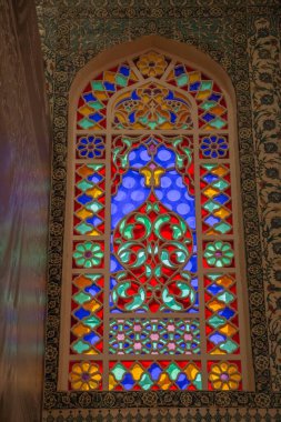 Göz kamaştırıcı, rengarenk cam vitrinler İslami geometrik desenler sergiliyor. Kırmızı, mavi ve yeşilin zengin mücevher tonları kemerli çerçeveyi doldurarak süslü bir zemin üzerinde büyüleyici bir ışık ve renk görüntüsü yaratıyor..