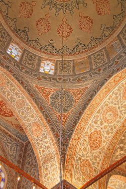 Düşük açılı bir görüntü, bir cami kubbesinin iç dekorasyonunu yakalar. Görsel özellikler karmaşık boyanmış tasarımlar, kemerli yapılar ve vitray pencereler.