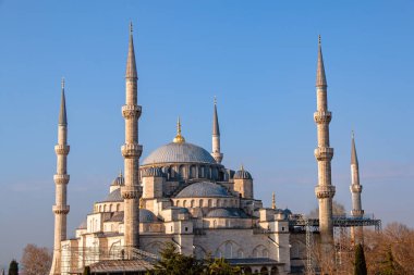 Türkiye 'nin İstanbul kentindeki Sultan Ahmed Camii' nin açık mavi bir gökyüzü altındaki görkemli manzarası. Fotoğrafta caminin altı minaresi, büyük merkezi kubbesi ve çevresindeki daha küçük kubbeler de dahil olmak üzere karmaşık ayrıntıları yer alıyor. Solgun taş 